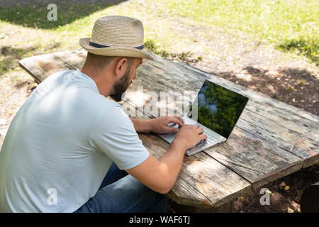 Man working on a laptop computer with a black screen for copy space, sitting on a bench in the summer outdoors in a park. back view Stock Photo
