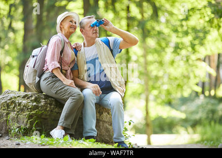 Senior man sitting and looking through binoculars with senior woman sitting near by him and smiling while they have a journey in the forest Stock Photo