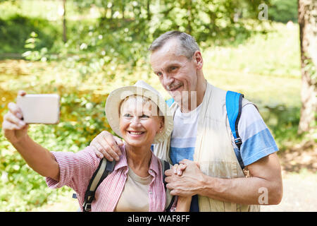 Happy mature woman standing and making selfie portrait with her husband on mobile phone while they standing in the park Stock Photo