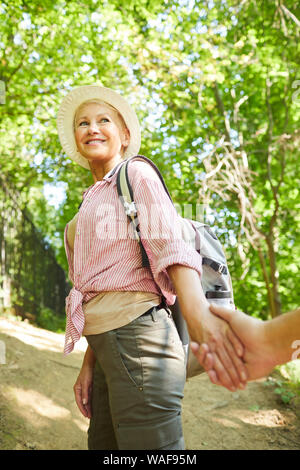 Happy mature woman in hat and with backpack behind her back holding hands with her man and smiling in nature outdoors Stock Photo