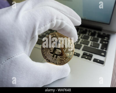 Men in white glove holding golden bitcoin against laptop. Focus on the coin. Stock Photo