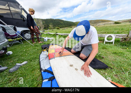 Surfer on vacation waxing surf board outdoors on the beach. Man is removing or applying wax to surfboard shortboard on vacation. Traces of wax are vis Stock Photo