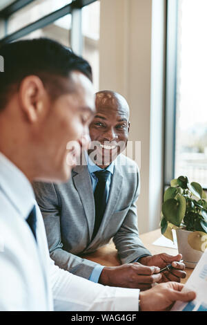 Smiling African American businessman discussing charts and graphs on paper with a colleague while sitting at an office table Stock Photo