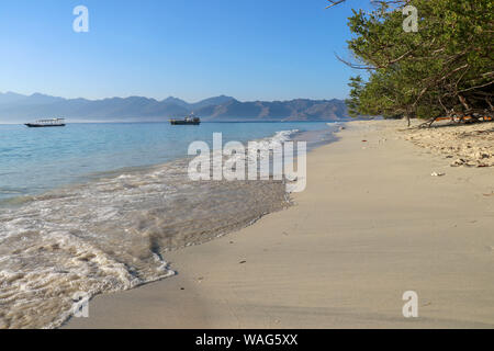 Sea water washes the fine yellow sand beach on Gili Meno Island in Indonesia. Mooring boat at sea not far from coast. Tropical shrubs and trees. Stock Photo