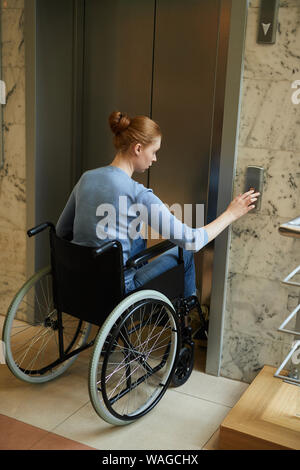 Young pretty woman sitting in wheelchair pushing the button and waiting for the elevator Stock Photo