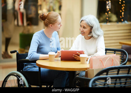 Senior mother talking to her disabled daughter in wheelchair and they using tablet pc while sitting at the table in cafe Stock Photo