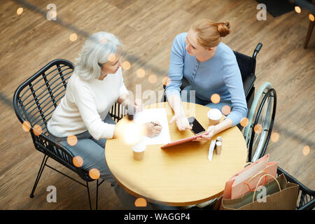 Young disabled woman pointing at digital tablet and explaining something to senior woman while she filling some document in cafe Stock Photo