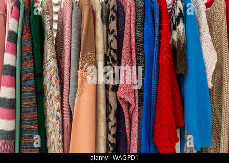 Row of knitted crocheted sweater and jumper hang on aluminium hanger clothes rack in retail fashion store or second hand outlet shop. Stock Photo