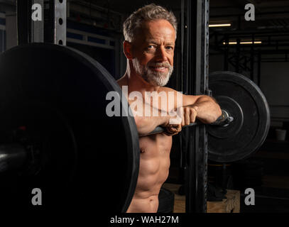 Black and white portrait of an attractive older male athlete with strong abs. The athletic and smiling bearded man is leaning against the barbell rack Stock Photo