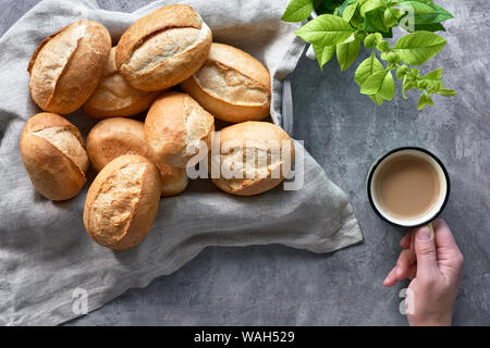 Bread buns in basket on rustic wood, spring leaves and hand with cup of coffee, top view on grey textured background Stock Photo