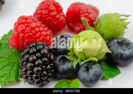 Mix of juicy forest berries and nuts: Raspberries, Blueberries, Blackberry with tiny green leaves and green hazelnuts. Close up, isolated on white. Stock Photo