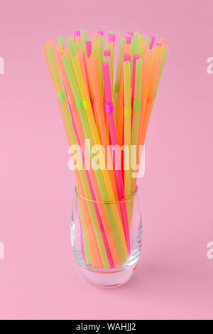 A lot of multicolored plastic cocktail tubes in a thick bottom glass on a pastel pink background. Drinking straws of bright neon colors. Front view. Stock Photo
