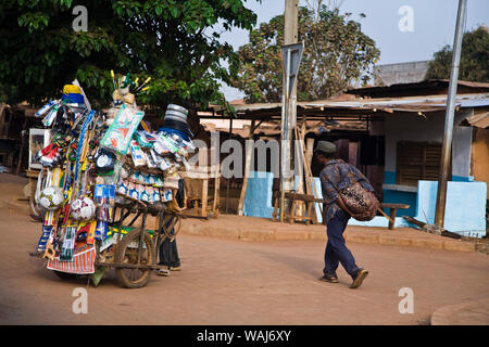 West Africa, Benin. Man walks down street, passing cart selling soccer balls, tools, and other goods. Stock Photo