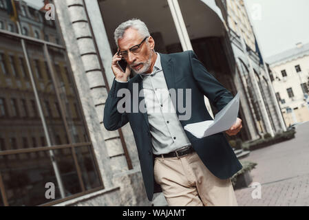 Senior businessman in suit holding papers and talking on the phone while walking outdoors. Business, technology, communication and people concept. Horizontal shot. Stock Photo
