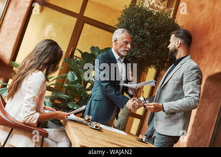 Portrait of three colleagues talking about their plans while sitting in a restaurant. Men are shaking hands while woman is looking through papers. Coffee cups on the table. Horizontal shot Stock Photo
