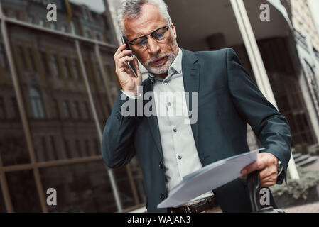 Senior businessman in suit holding papers and talking on the phone while walking outdoors. Business, technology, communication and people concept. Horizontal shot. Stock Photo