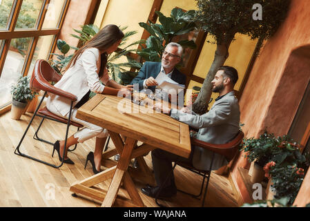 Cropped portrait of three colleagues talking about their plans while sitting in a restaurant. Coffee cups on the table. Horizontal shot. Dutch angle Stock Photo