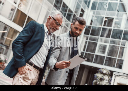Portrait of senior and young businessmen in suits talking about their plans outdoors. Young colleague shows his ideas using tablet pc. Horizontal shot Stock Photo