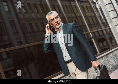 Senior businessman with briefcase talking on the phone while walking outdoors. Business, technology, communication and people concept. Horizontal shot. Stock Photo