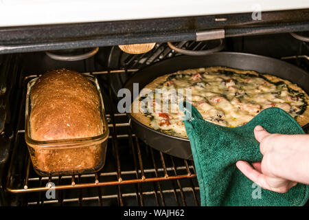 Woman removing homemade vegetarian pizza from oven. Also in the oven is a homemade multi-grain loaf of bread made with the same dough as the pizza crust. (MR) Stock Photo