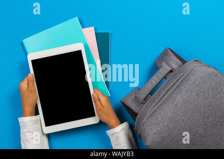 Kid hands with tablet computer, notebooks, books and gray backpack over light sky blue background Stock Photo