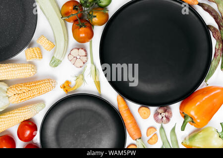 Pattern made of autumn vegetables. Tomatoes, onion, carrot, garlic, red beet, pepper, zucchini, corn, green haricot and black plate on white backgroun Stock Photo
