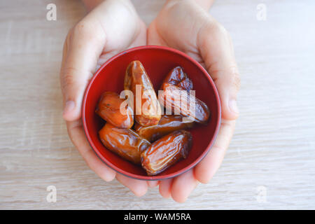 hand holding red bowl with dried fruits from date palm on wooden background, phoenix dactylifera on red bowl in hand. Stock Photo