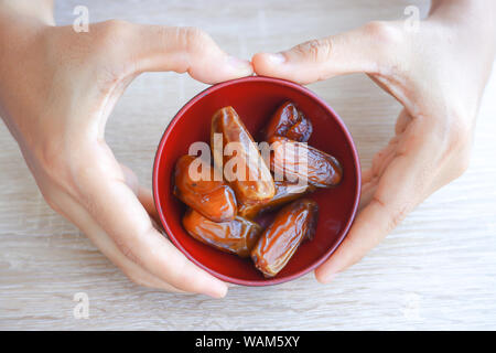 hand holding red bowl with dried fruits from date palm on wooden background, phoenix dactylifera on red bowl in hand. Stock Photo