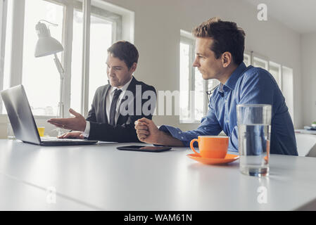 Two businessmen sitting at an office desk working together on a project using laptop and digital tablet. Stock Photo