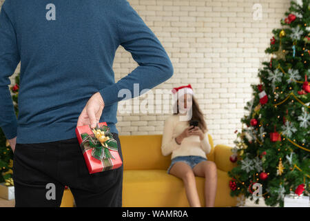 Man wearing blue sweater holding hid red gift box on the back is congratulating girlfriend and giving her gift in christmas festival on  blur backgrou Stock Photo