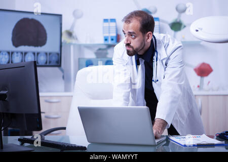 Portrait of doctor working on laptop and looking over his monitor. Doctor on his cabinet. Doctor in a laboratory.