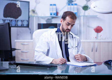 Young and smart doctor checking his patient list on his clipboard. Handsome doctor with wite coat and stethoscope. Stock Photo
