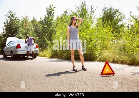 Young multiethnic couple broke down the car while traveling on the way to rest. They are trying to fix the broken by their own or should hitchhike, getting nervous. Relationship, troubles on the road, vacation. Stock Photo