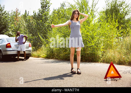 Young multiethnic couple broke down the car while traveling on the way to rest. They are trying to fix the broken by their own or should hitchhike, getting nervous. Relationship, troubles on the road, vacation. Stock Photo