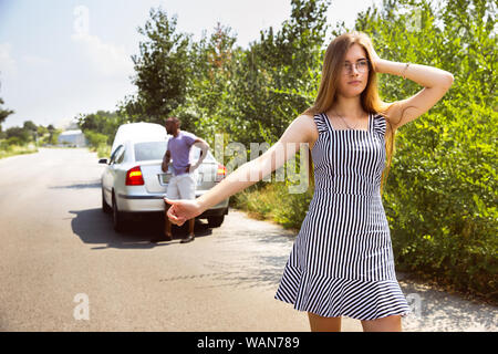 Young multiethnic couple broke down the car while traveling on the way to rest. They are trying to fix the broken by their own or should hitchhike, getting nervous. Relationship, troubles on the road, vacation. Stock Photo