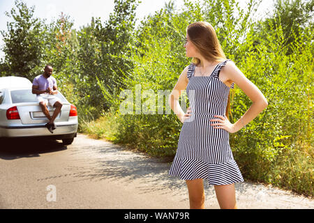 Young multiethnic couple broke down the car while traveling on the way to rest. They are trying to fix the broken by their own or should hitchhike, getting nervous. Relationship, troubles on the road, vacation. Stock Photo