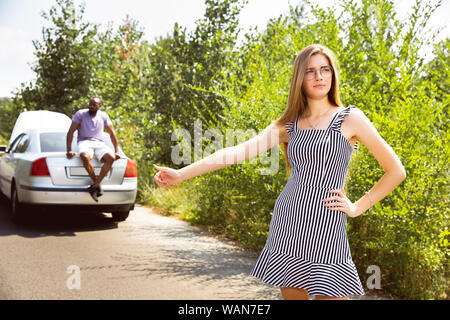 Young multiethnic couple broke down the car while traveling on the way to rest. They are trying to fix the broken by their own or should hitchhike, getting nervous. Relationship, troubles on the road, vacation. Stock Photo