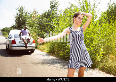 Young multiethnic couple broke down the car while traveling on the way to rest. They are trying to fix the broken by their own or should hitchhike, getting nervous. Relationship, troubles on the road, vacation. Stock Photo