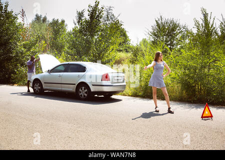 Young multiethnic couple broke down the car while traveling on the way to rest. They are trying to fix the broken by their own or should hitchhike, getting nervous. Relationship, troubles on the road, vacation. Stock Photo