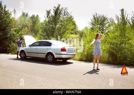 Young multiethnic couple broke down the car while traveling on the way to rest. They are trying to fix the broken by their own or should hitchhike, getting nervous. Relationship, troubles on the road, vacation. Stock Photo