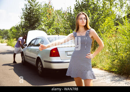 Young multiethnic couple broke down the car while traveling on the way to rest. They are trying to fix the broken by their own or should hitchhike, getting nervous. Relationship, troubles on the road, vacation. Stock Photo