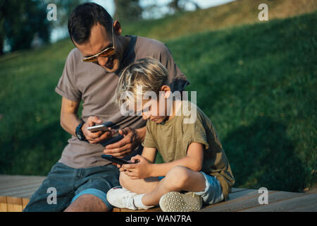 Photo of young man and boy with phones in hands sitting behind wooden fence in park on summer day Stock Photo