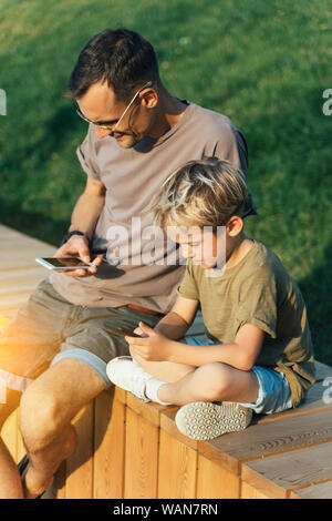 Photo of man and boy with phones in hands sitting behind wooden fence in park on summer day Stock Photo