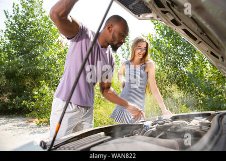 Young multiethnic couple broke down the car while traveling on the way to rest. They are trying to fix the broken by their own or should hitchhike, getting nervous. Relationship, troubles on the road, vacation. Stock Photo