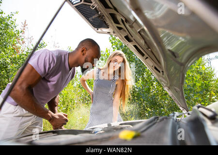 Young multiethnic couple broke down the car while traveling on the way to rest. They are trying to fix the broken by their own or should hitchhike, getting nervous. Relationship, troubles on the road, vacation. Stock Photo