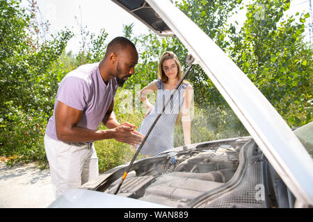 Young multiethnic couple broke down the car while traveling on the way to rest. They are trying to fix the broken by their own or should hitchhike, getting nervous. Relationship, troubles on the road, vacation. Stock Photo