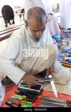 Man in traditional clothes selling knives, sharpening a knife at Sinaw market, Oman Stock Photo