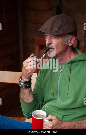 A middle aged man sitting in a shed and smoking a pipe. Stock Photo