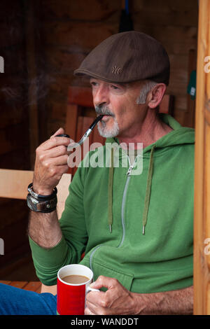 A middle aged man sitting in a shed and smoking a pipe. Stock Photo
