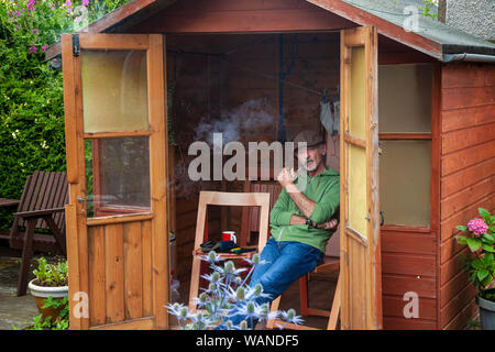 A middle aged man sitting in a garden shed and smoking a pipe. Stock Photo